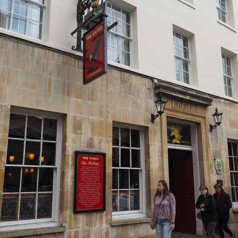 Exterior of The Eagle pub in Cambridge, with its hanging sign, stone facade, red doorway and three pedestrians walking past.