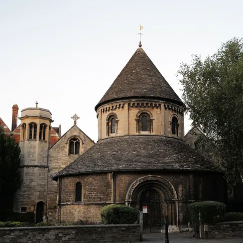 The Round Church in Cambridge, a circular Norman stone church built in 1130, seen from across the street at dusk beside trees.