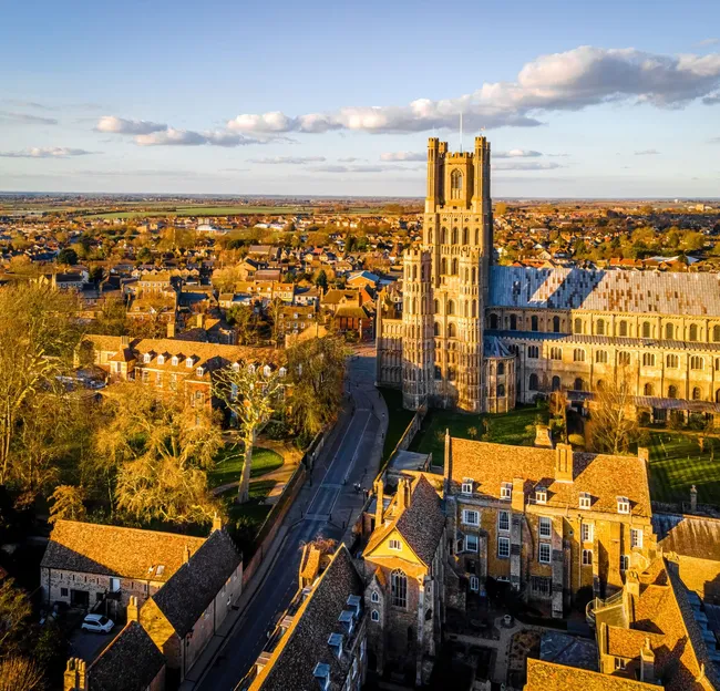 Aerial view of Ely, Cambridgeshire, with Ely Cathedral glowing in golden evening light above the surrounding town and open countryside.