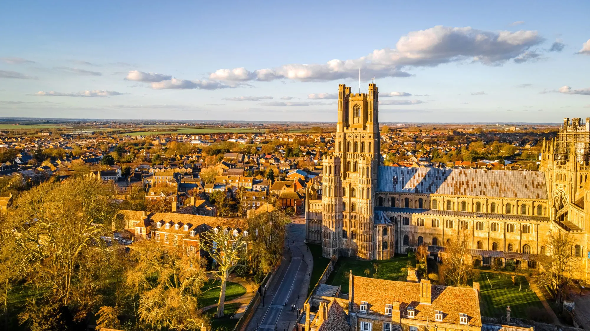 Aerial view of Ely, Cambridgeshire, with Ely Cathedral glowing in golden evening light above the surrounding town and open countryside.