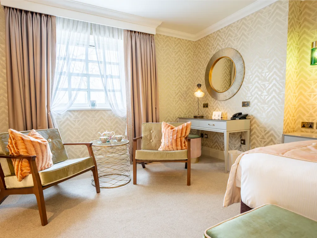 Elegant hotel bedroom with two upholstered armchairs, a small round table, a desk and soft blush curtains by the window