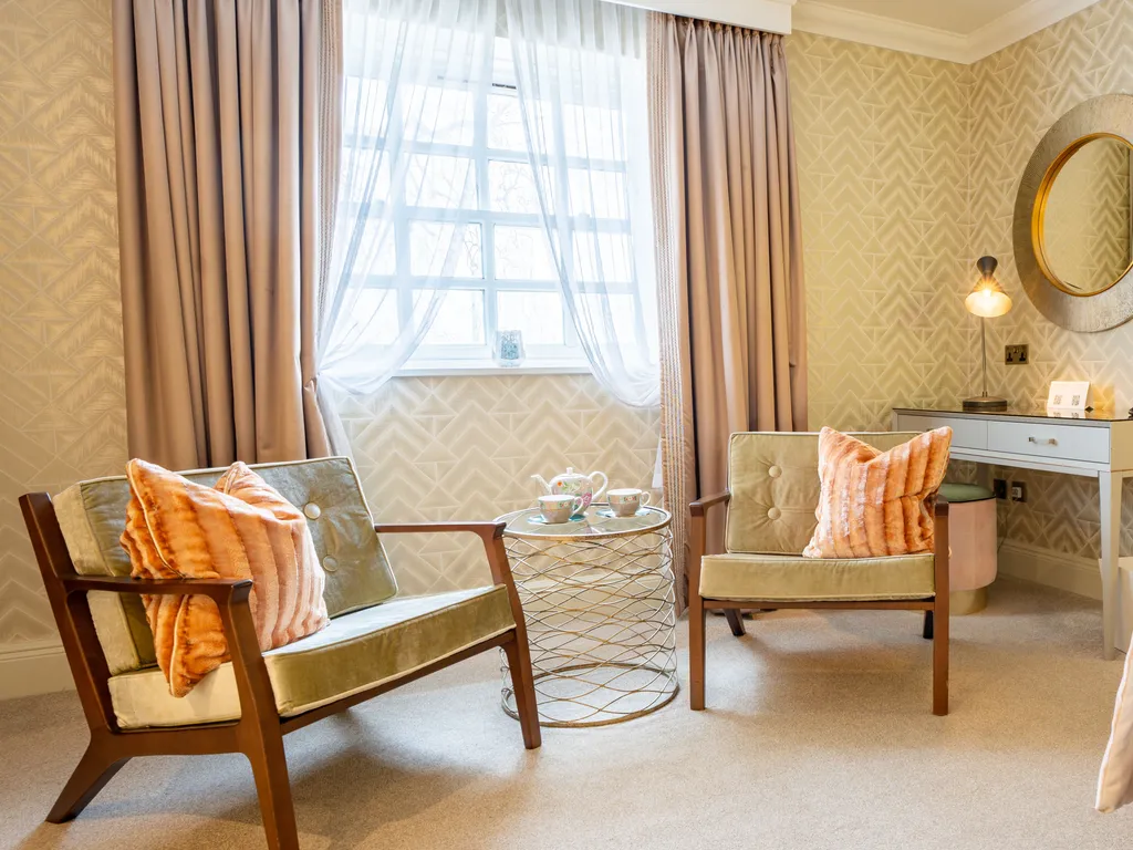 Elegant hotel bedroom with two upholstered armchairs, a small round table, a desk and soft blush curtains by the window