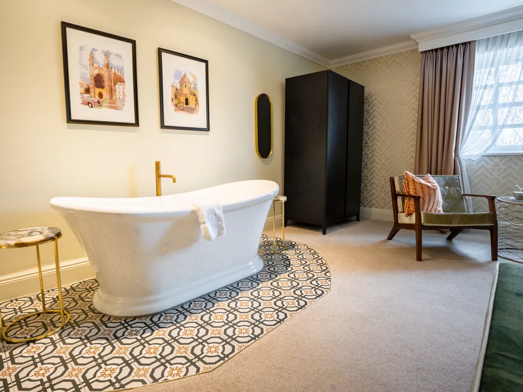 Elegant bathroom with a freestanding white bath on patterned floor tiles, framed artwork, a black wardrobe, and a seating area by the window