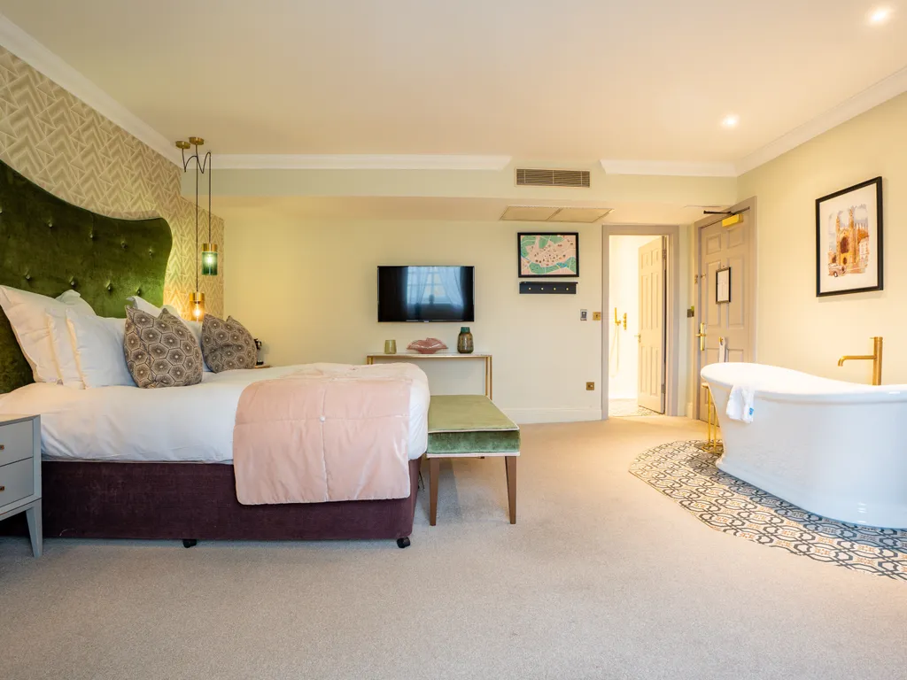 Spacious boutique hotel bedroom with a green upholstered headboard, wall-mounted television and freestanding bath
