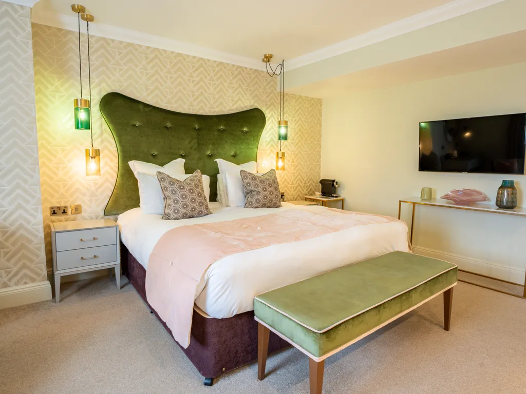 Stylish bedroom with a large green upholstered headboard, pendant lights, wall-mounted television and matching bench at the foot of the bed