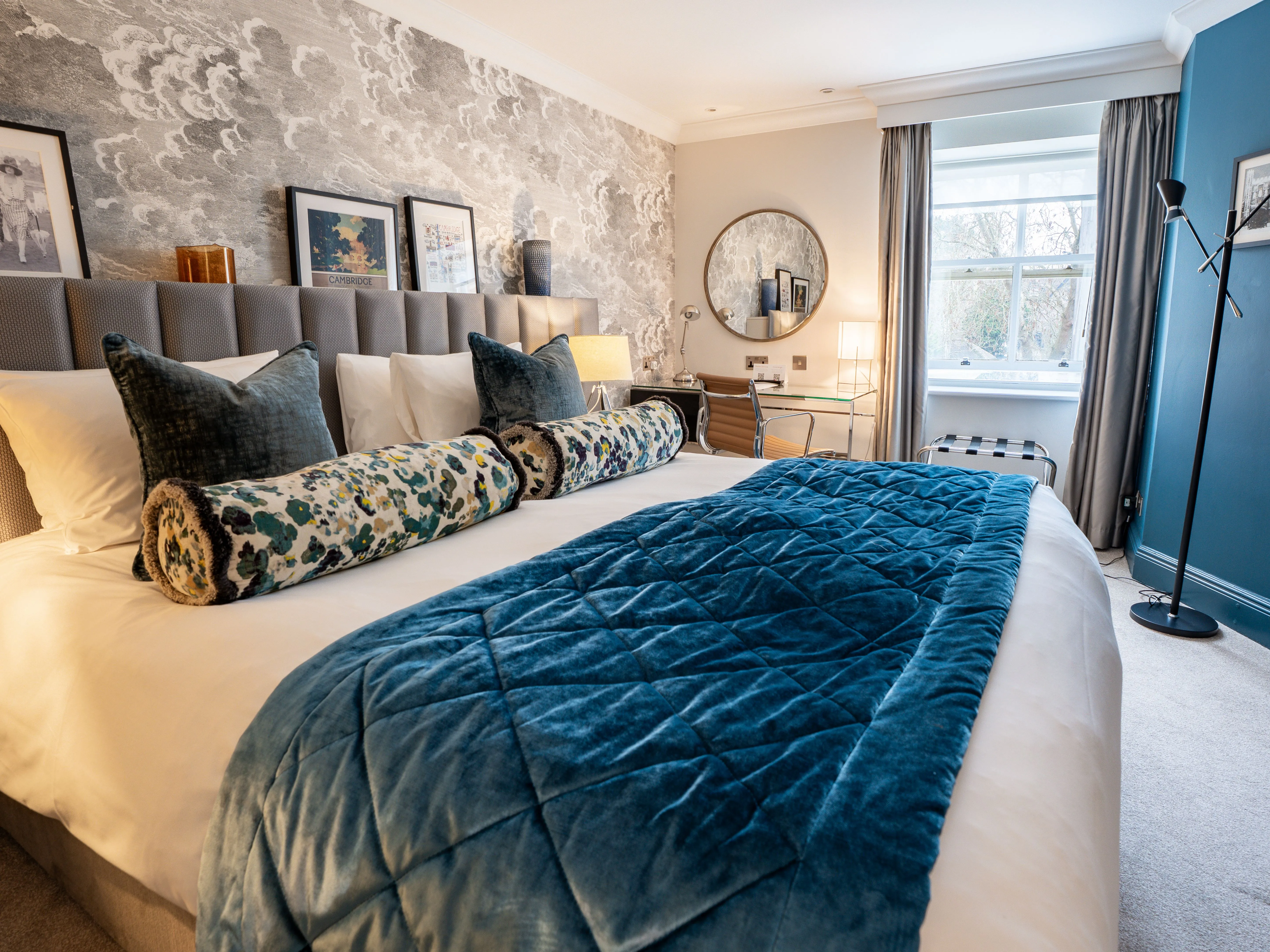 Elegant bedroom with a large upholstered bed, blue velvet throw, patterned wallpaper and a desk by the window