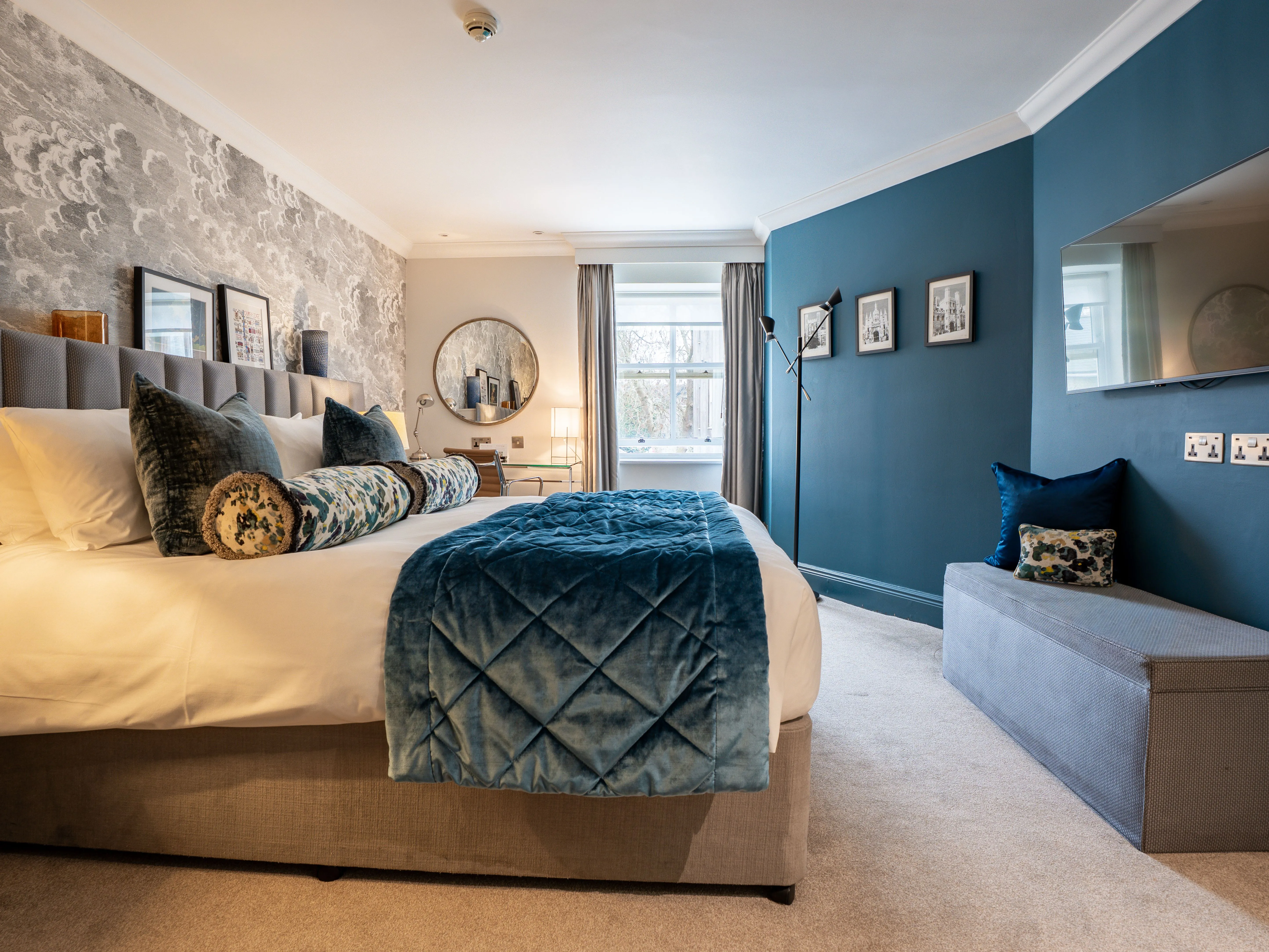 Elegant bedroom with a large upholstered bed, blue accent wall, wall-mounted television and window seating bench