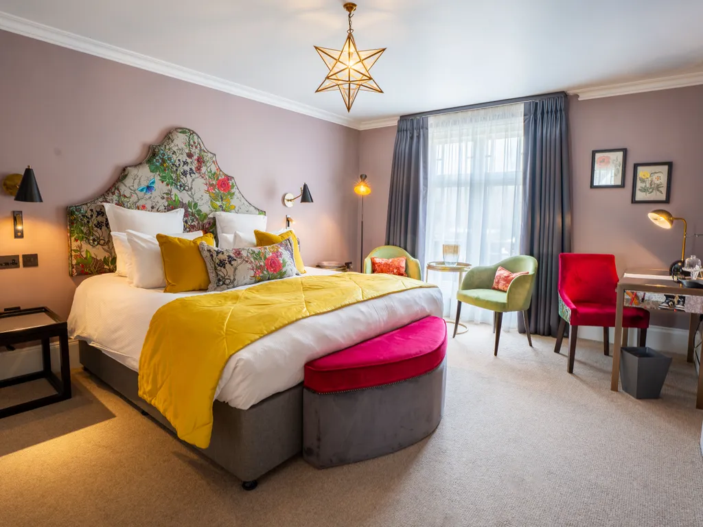 Stylish hotel bedroom with a floral upholstered headboard, mustard bed runner, pink bench and seating area by the window