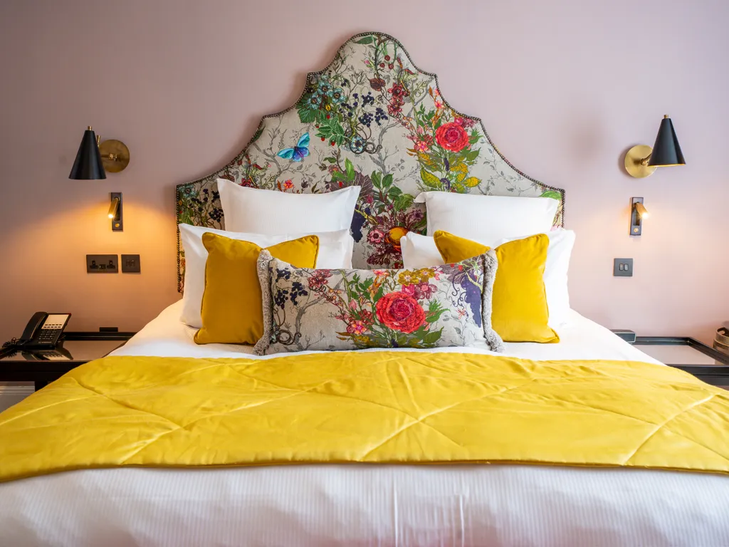 Stylish hotel bedroom with a floral upholstered headboard, mustard yellow cushions and throw, and soft pink walls