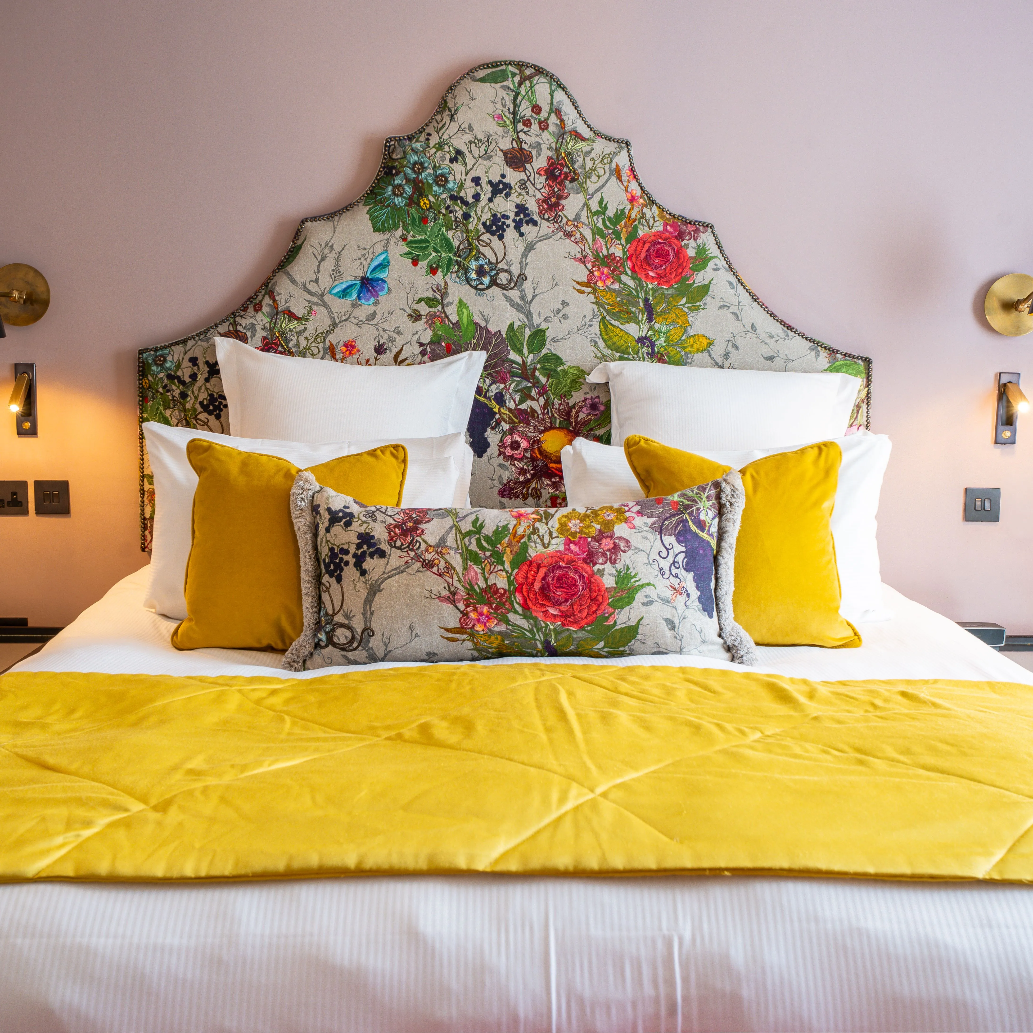 Stylish hotel bedroom with a floral upholstered headboard, mustard yellow cushions and throw, and soft pink walls