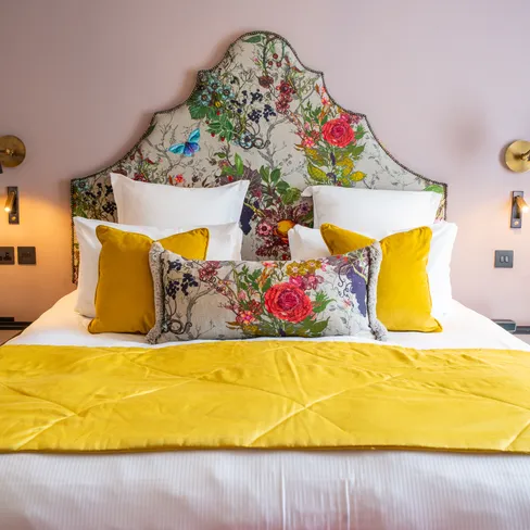 Stylish hotel bedroom with a floral upholstered headboard, mustard yellow cushions and throw, and soft pink walls