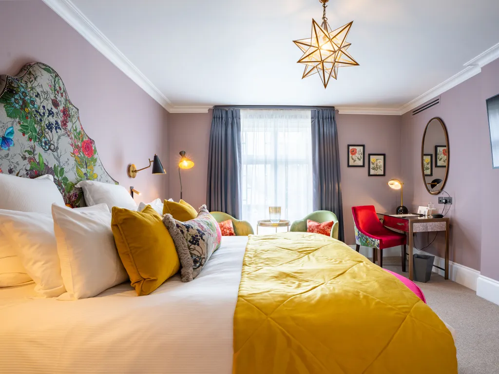 Stylish boutique bedroom with a floral upholstered headboard, mustard bed runner, pastel walls and a dressing table beneath a round mirror
