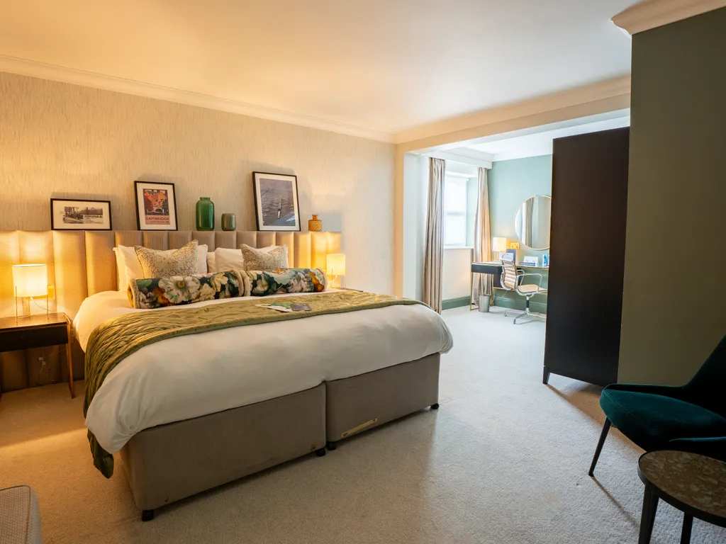 Spacious hotel bedroom with a large bed, bedside lamps, framed artwork and a dressing desk by the window