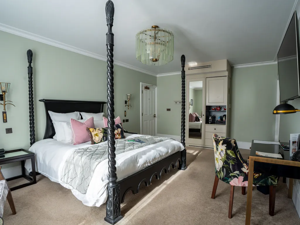 Elegant bedroom with a dark four-poster bed, soft green walls, floral chairs and a desk with a wall-mounted television