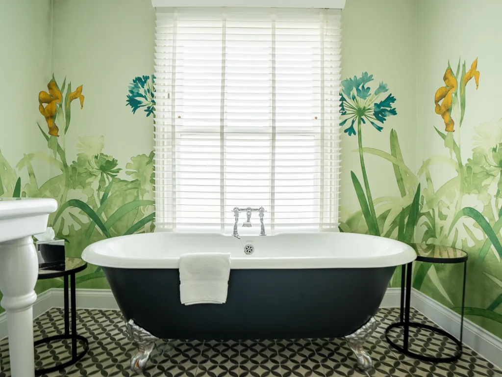 Freestanding black and white clawfoot bath beneath a window with white blinds, framed by botanical mural wallpaper in a bright bathroom