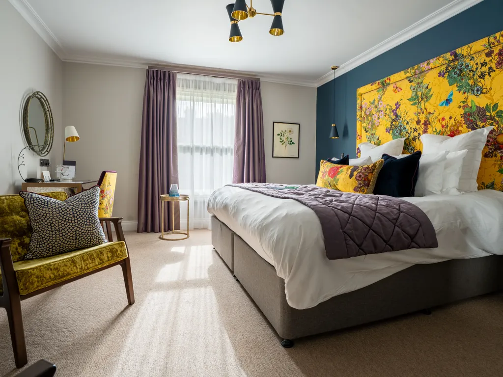 Stylish bedroom with a large bed, bright floral headboard, dark teal feature wall, purple curtains and a velvet armchair