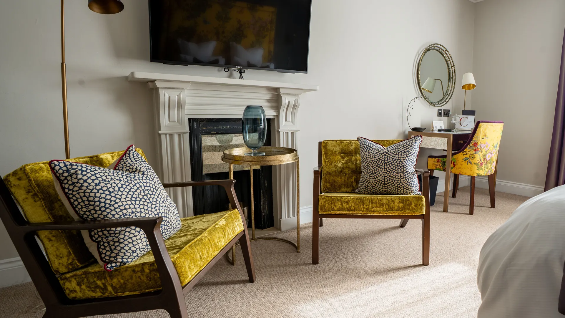 Elegant bedroom seating area with mustard velvet armchairs, a fireplace, wall-mounted television and purple curtains