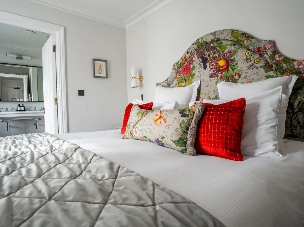Elegant bedroom with a floral upholstered headboard, white bedding, red accent cushions and an en-suite bathroom visible through an open door