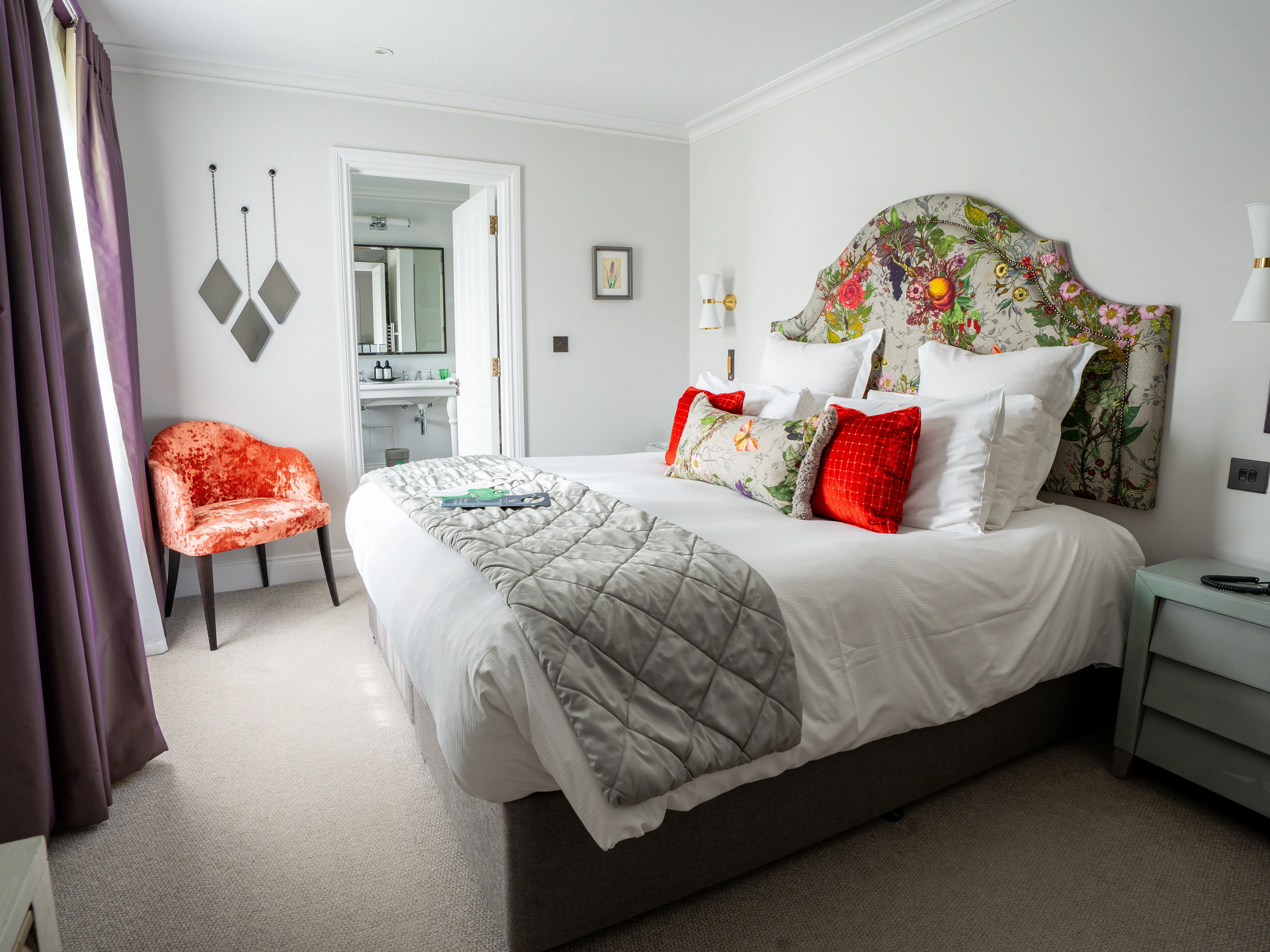 Elegant hotel bedroom with a floral upholstered headboard, white bedding, red cushions and an en-suite bathroom