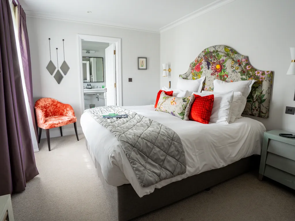Elegant hotel bedroom with a floral upholstered headboard, white bedding, red cushions and an en-suite bathroom
