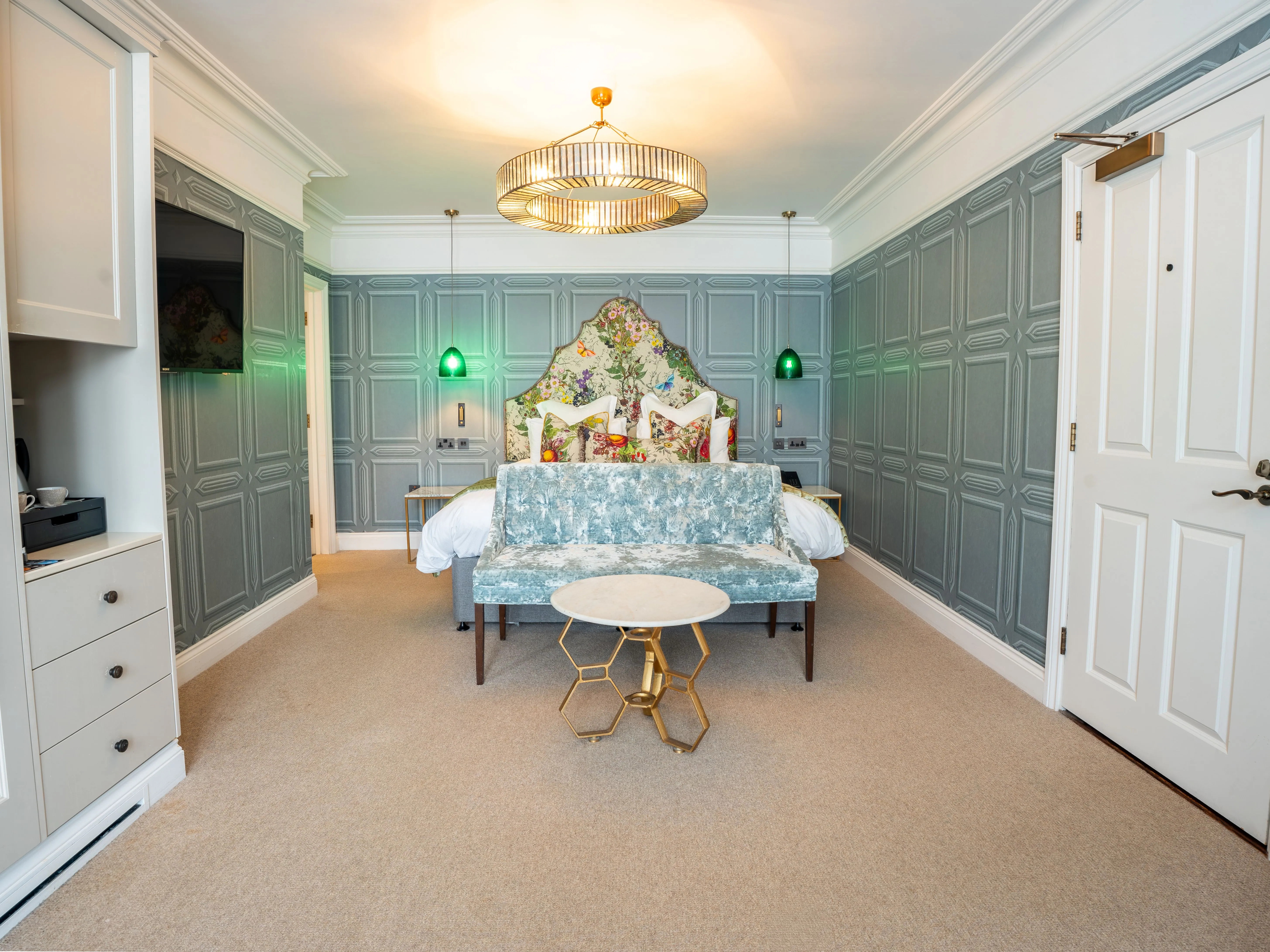 Elegant boutique-style bedroom with blue-grey panelled walls, floral headboard, velvet bench and gold chandelier