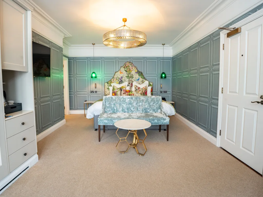 Elegant boutique-style bedroom with blue-grey panelled walls, floral headboard, velvet bench and gold chandelier