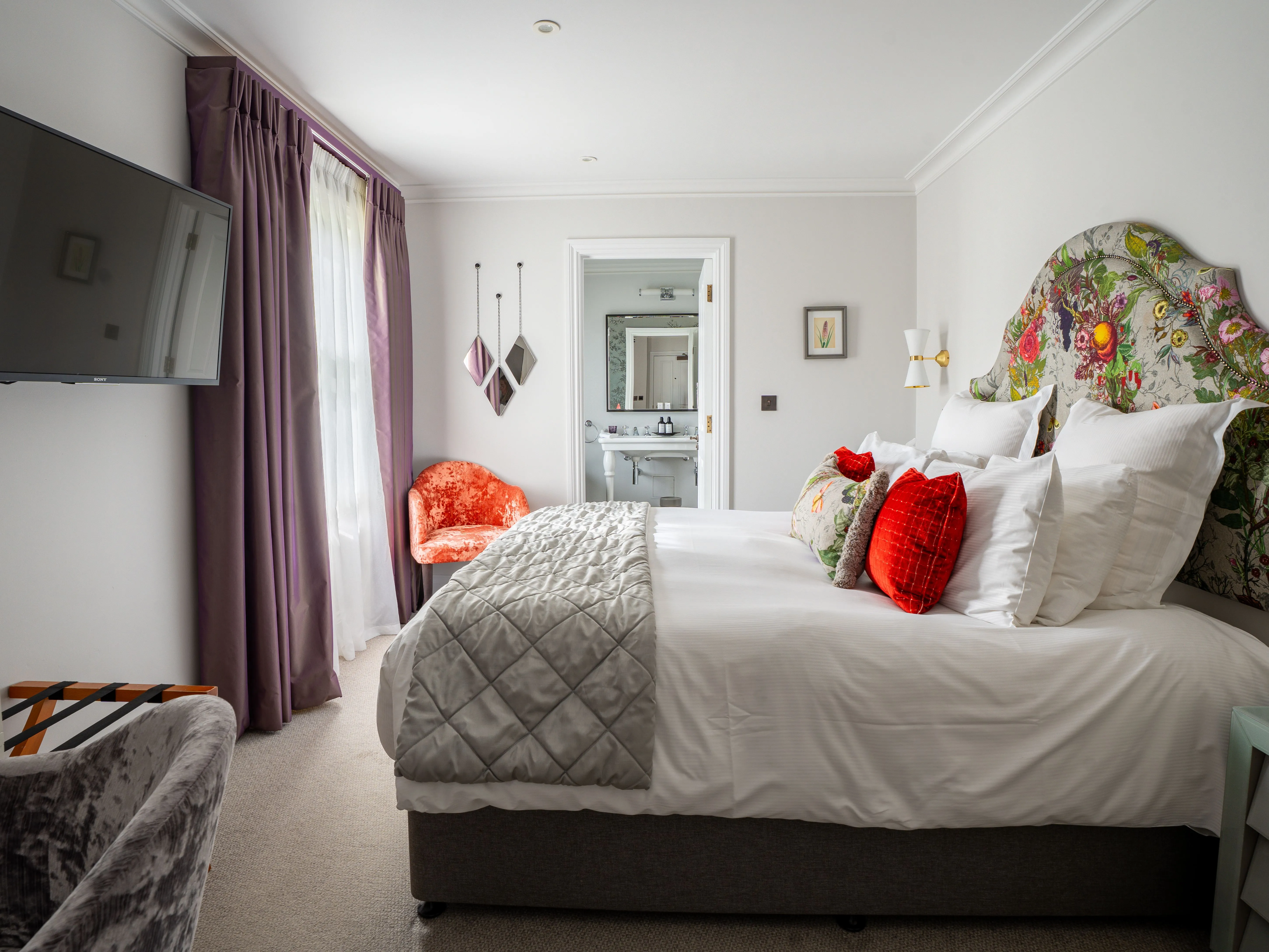Elegant bedroom with floral upholstered headboard, white bedding, purple curtains, wall-mounted television and en-suite bathroom