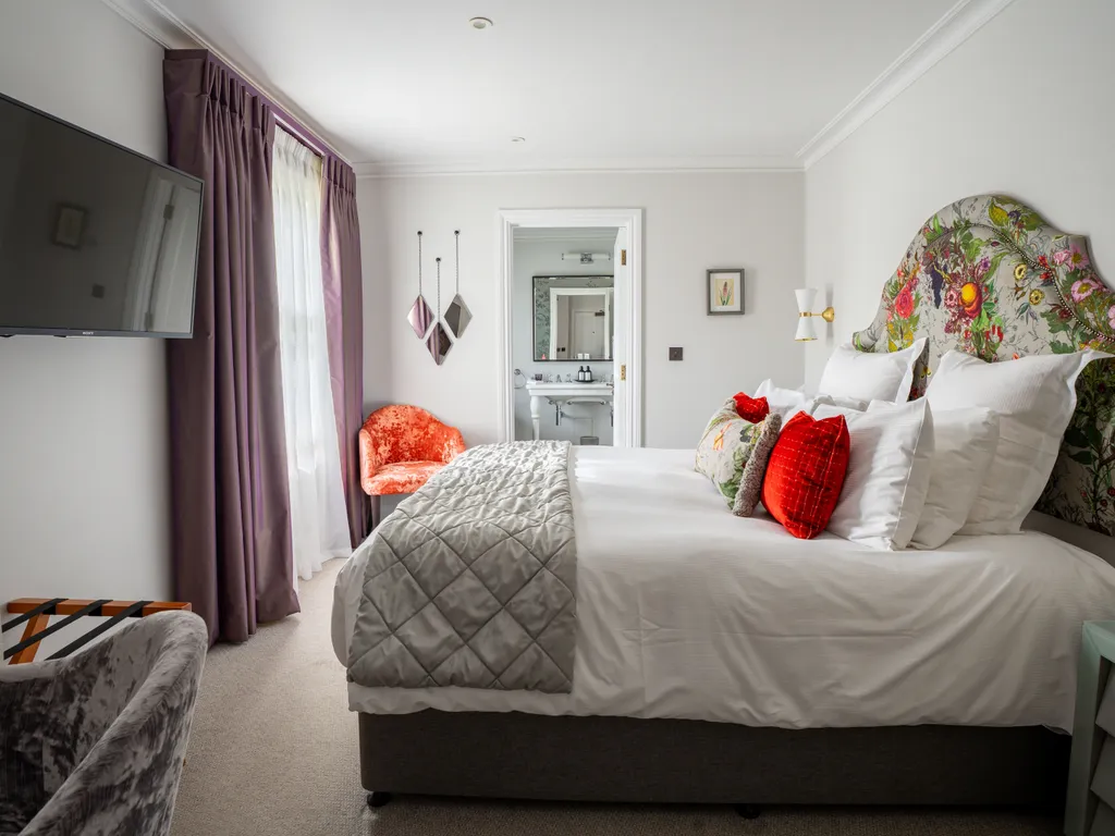 Elegant bedroom with floral upholstered headboard, white bedding, purple curtains, wall-mounted television and en-suite bathroom