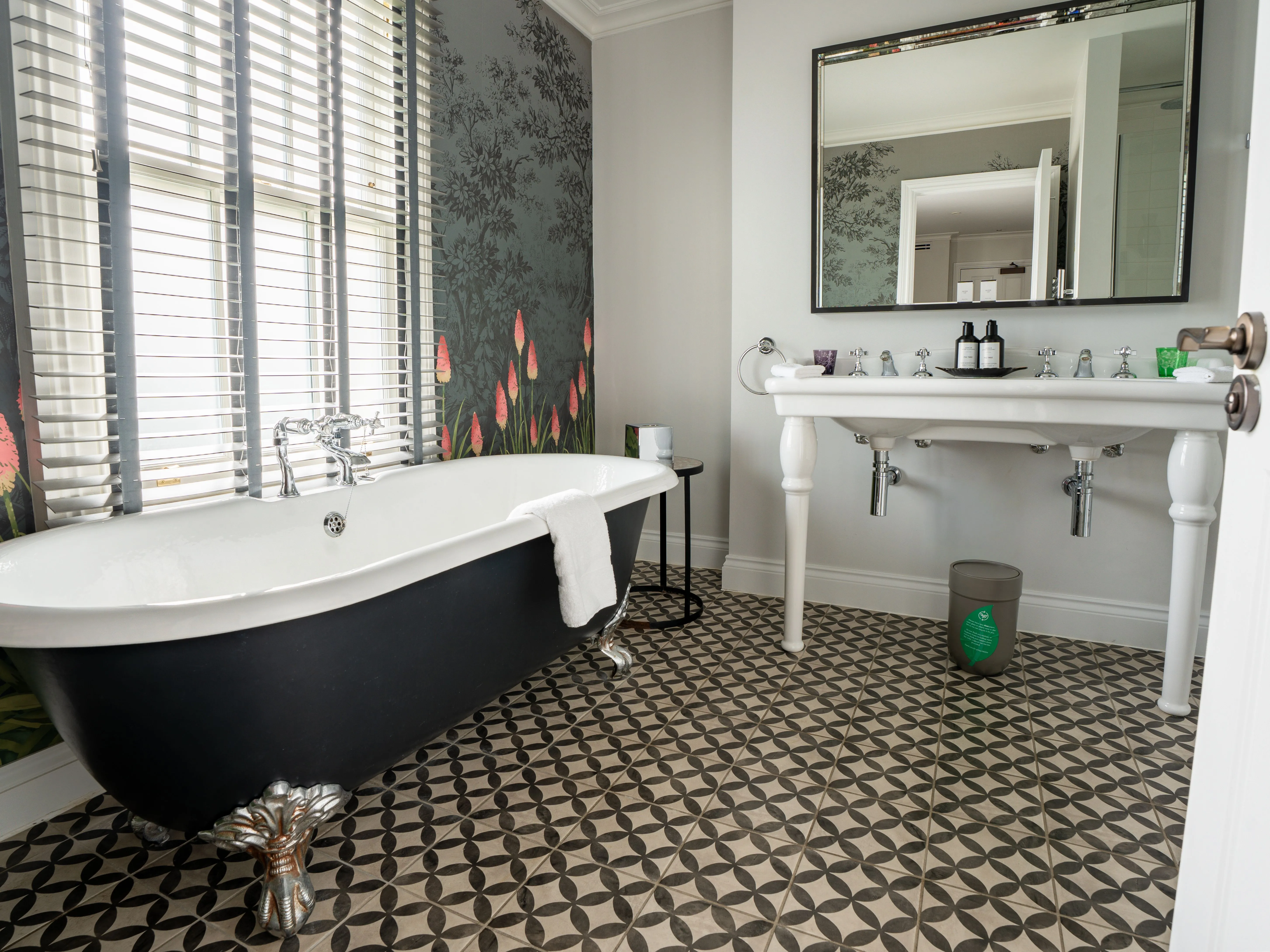 Elegant bathroom with a black freestanding roll-top bath, patterned floor tiles, floral wallpaper and a white double sink beneath a large mirror.