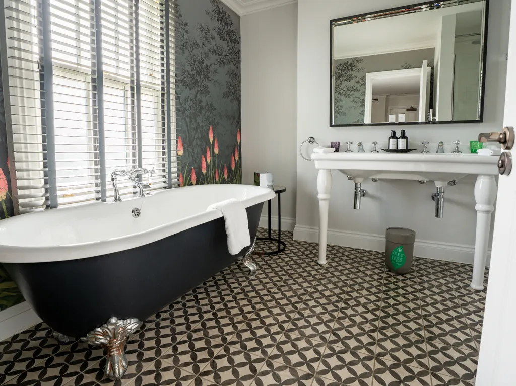 Elegant bathroom with a black freestanding roll-top bath, patterned floor tiles, floral wallpaper and a white double sink beneath a large mirror.