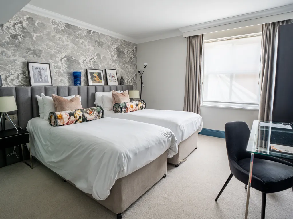 Modern twin hotel room with patterned feature wall, two single beds, bedside tables and a glass desk by the window
