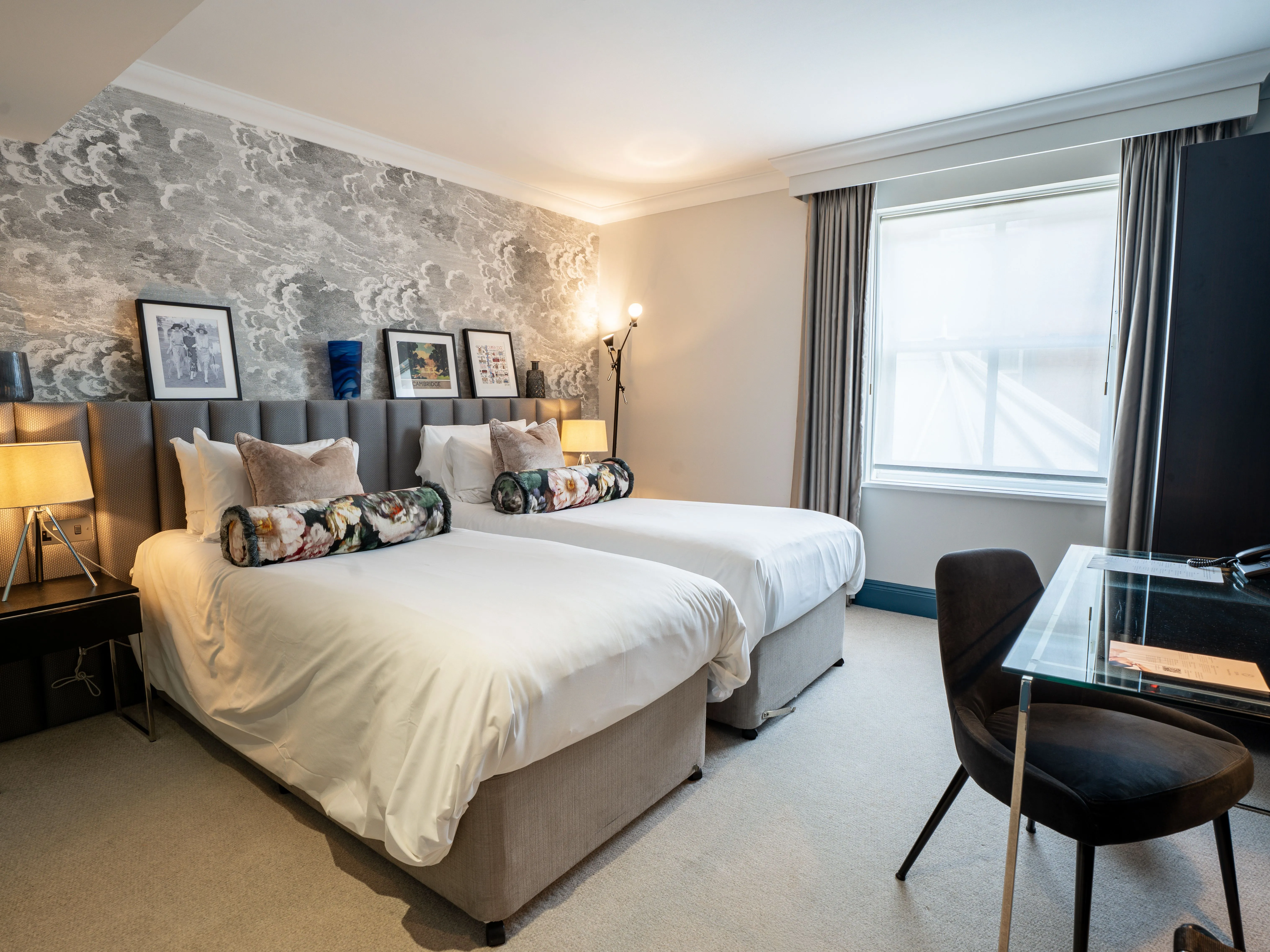 Modern twin hotel room with two single beds, patterned feature wall, bedside lamps and a glass desk by the window