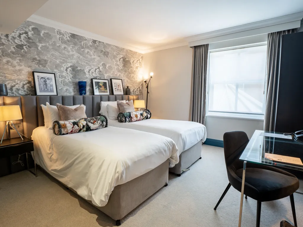 Modern twin hotel room with two single beds, patterned feature wall, bedside lamps and a glass desk by the window