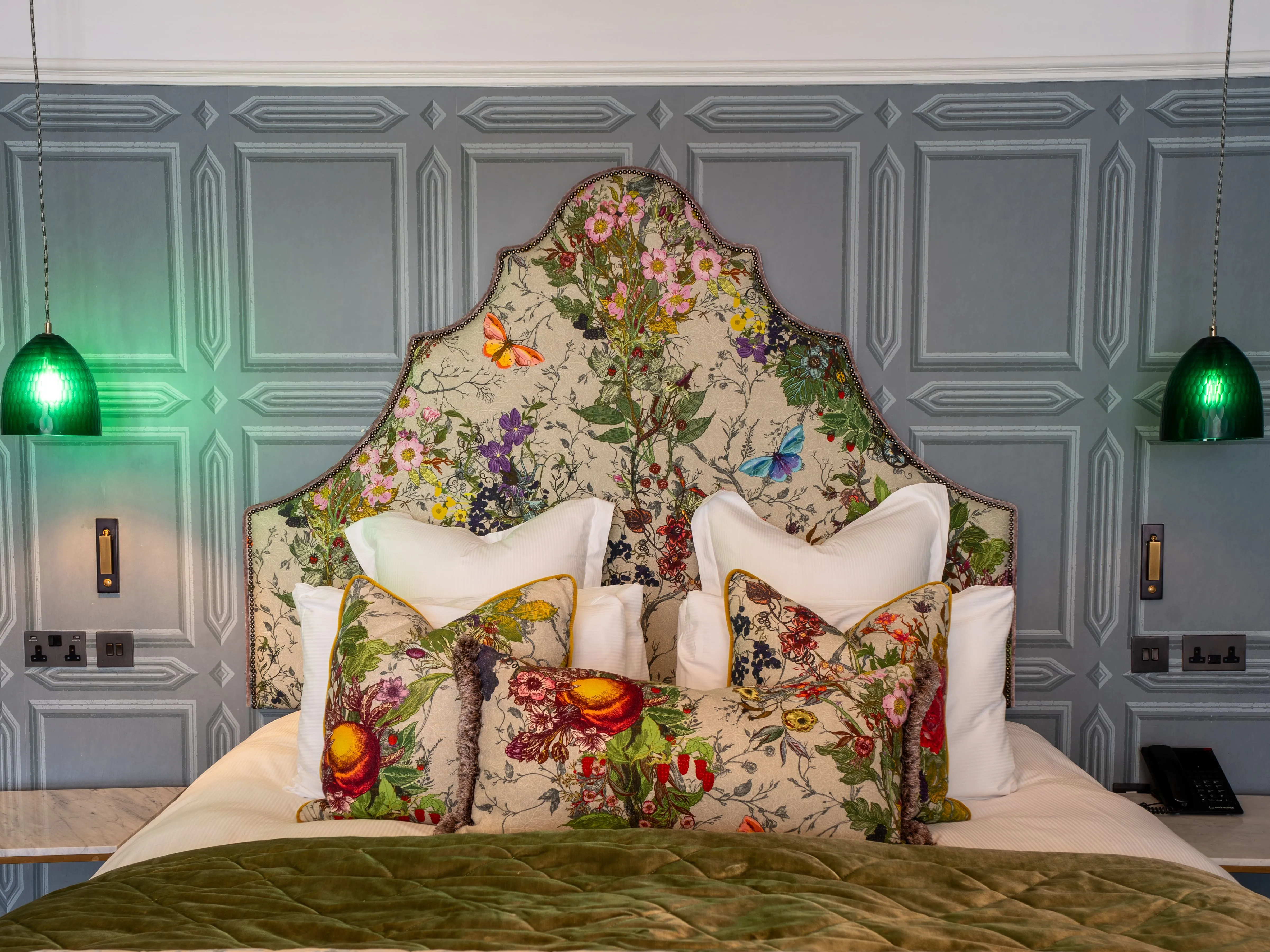 Elegant bedroom with a floral upholstered headboard, matching cushions, grey panelled walls and green pendant lights