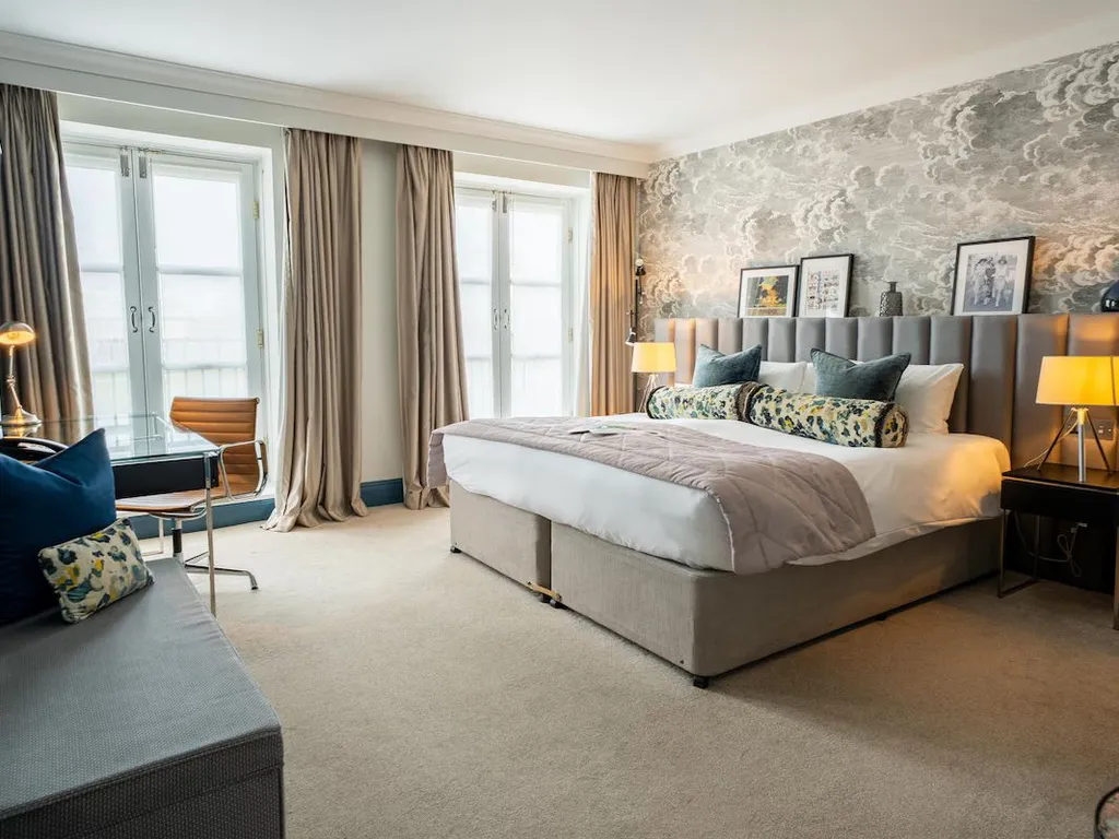 Spacious boutique hotel bedroom with a large upholstered bed, patterned wallpaper, bedside lamps, desk and tall windows with curtains.