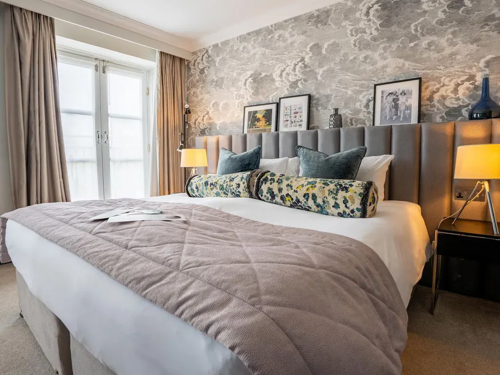 Elegant bedroom with a large upholstered bed, patterned bolster cushions, bedside lamps and cloud-effect wallpaper