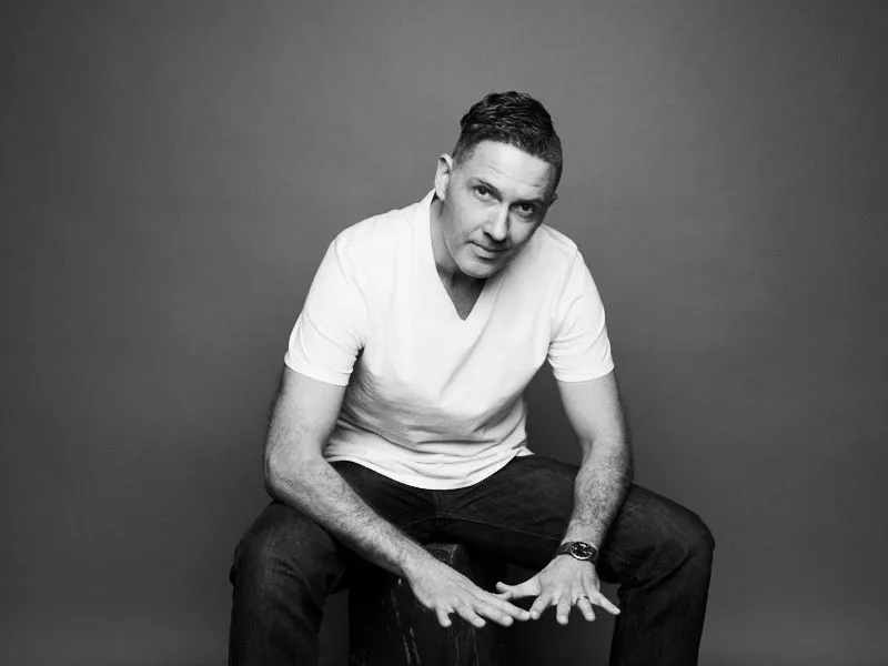 Black and white studio portrait of a man seated on a wooden box, wearing a white T-shirt, dark jeans and trainers.