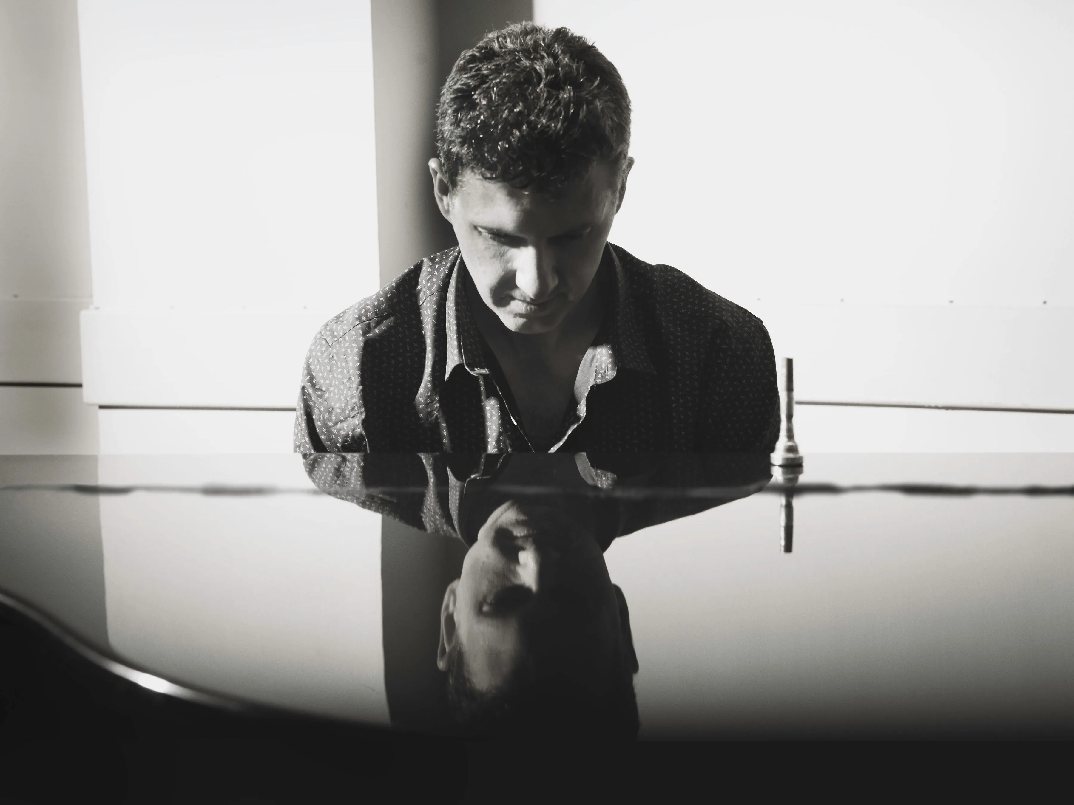 Black and white portrait of a man leaning over a grand piano with his reflection visible on the glossy surface