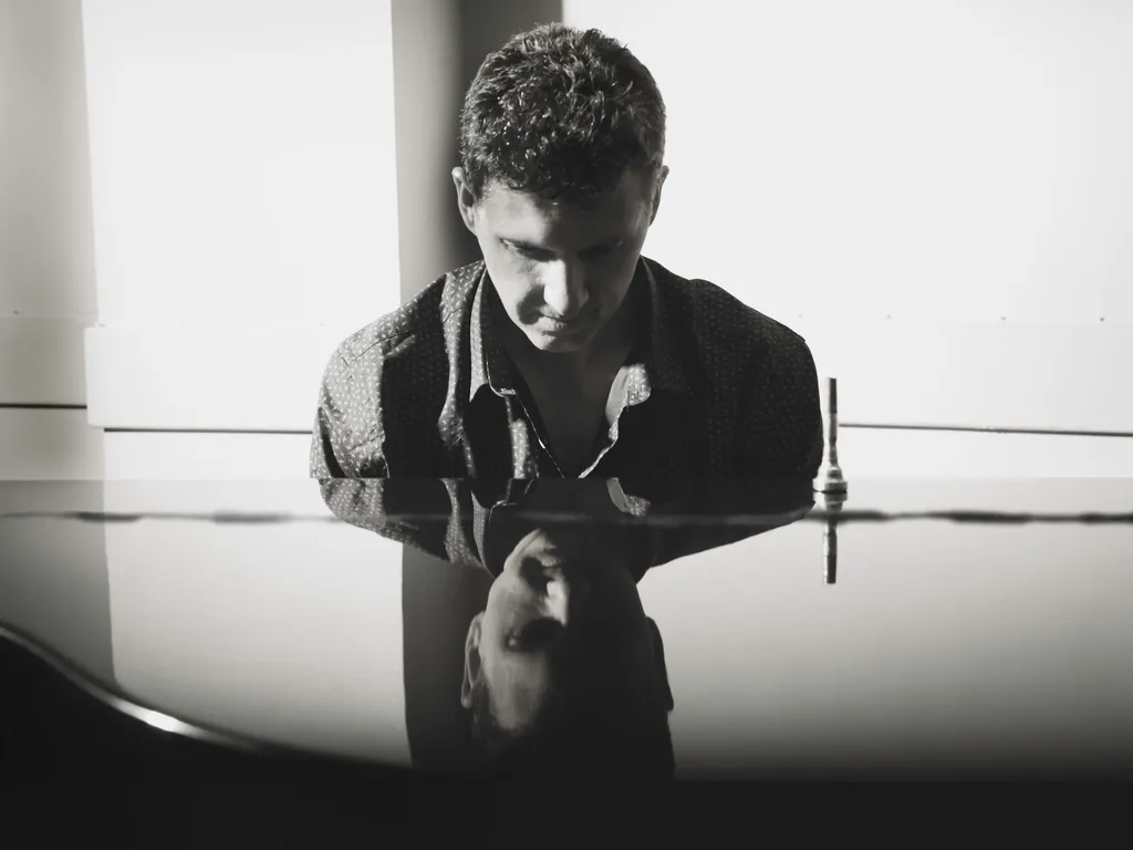 Black and white portrait of a man leaning over a grand piano with his reflection visible on the glossy surface