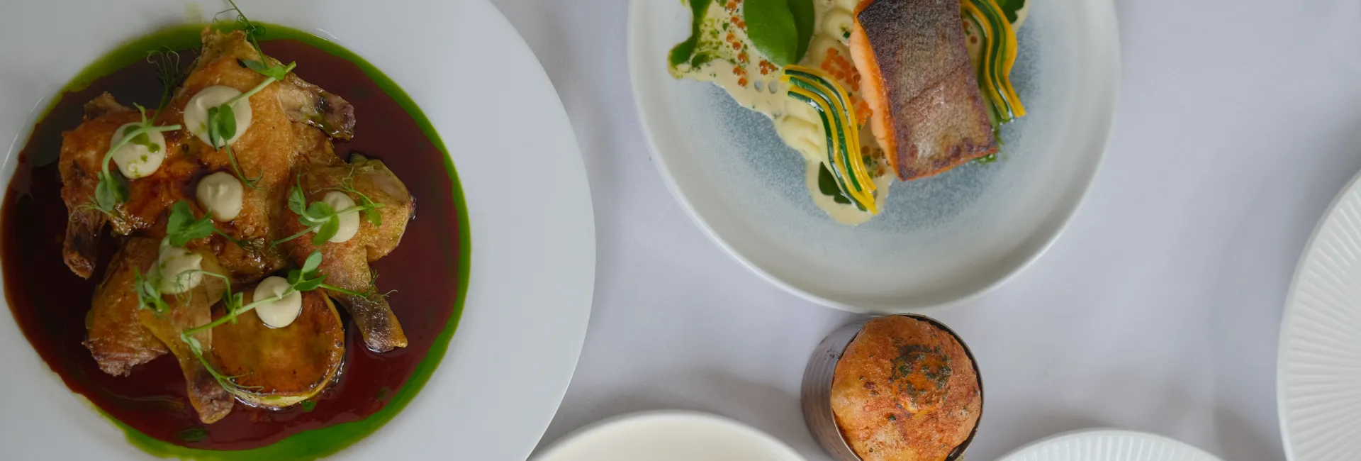 Overhead view of elegantly plated gourmet dishes on a white tablecloth, including roasted poultry, seared fish and a vibrant starter.