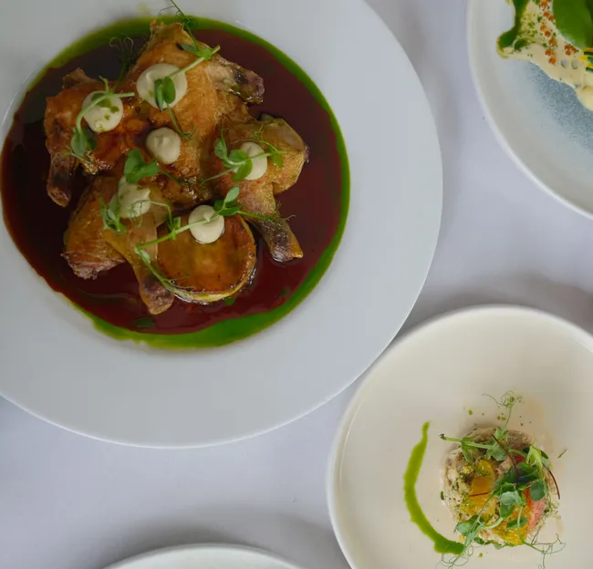 Overhead view of elegantly plated gourmet dishes on a white tablecloth, including roasted poultry, seared fish and a vibrant starter.