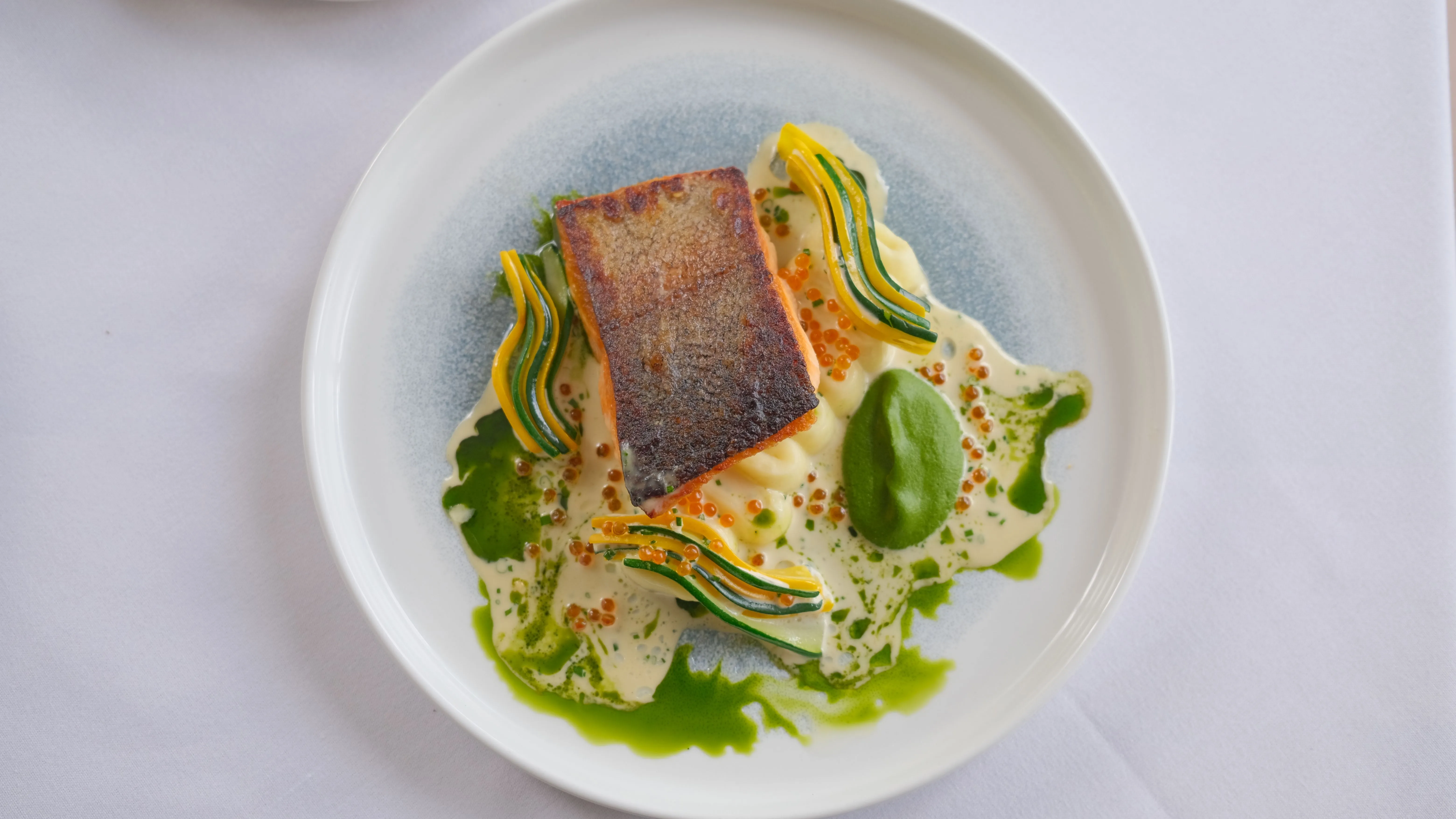 Pan-seared salmon fillet served on a white plate with a creamy sauce, green herb oil, fish roe and thin ribbons of courgette and yellow squash.