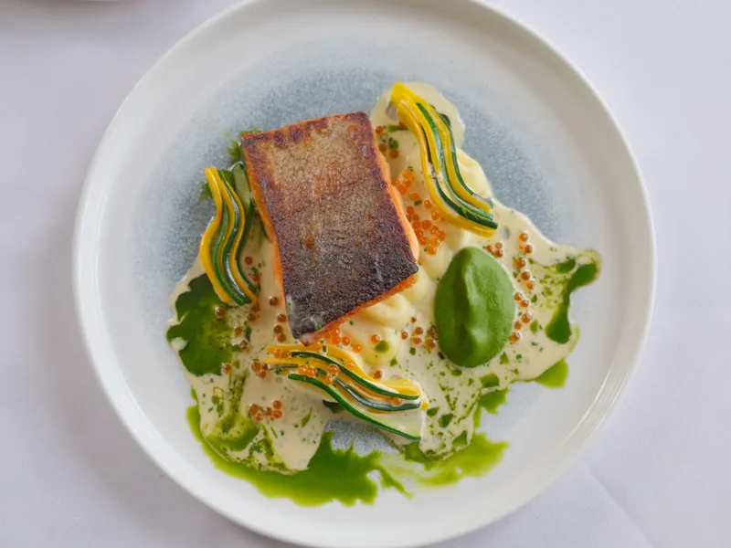 Pan-seared salmon fillet served on a white plate with a creamy sauce, green herb oil, fish roe and thin ribbons of courgette and yellow squash.