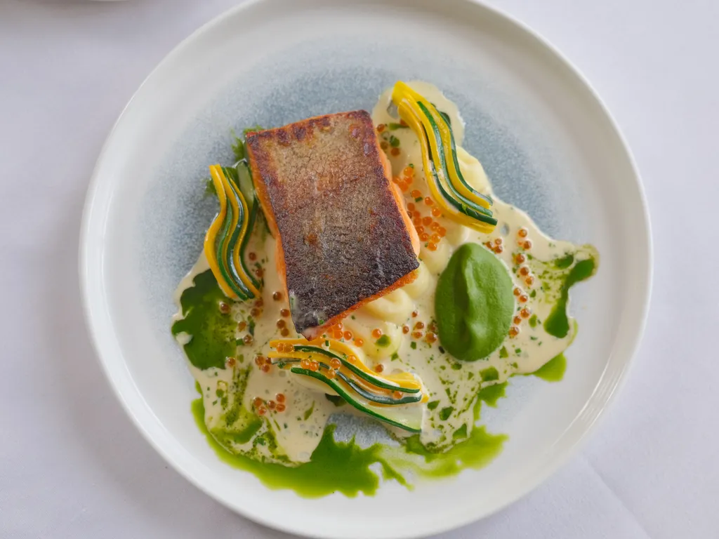 Pan-seared salmon fillet served on a white plate with a creamy sauce, green herb oil, fish roe and thin ribbons of courgette and yellow squash.