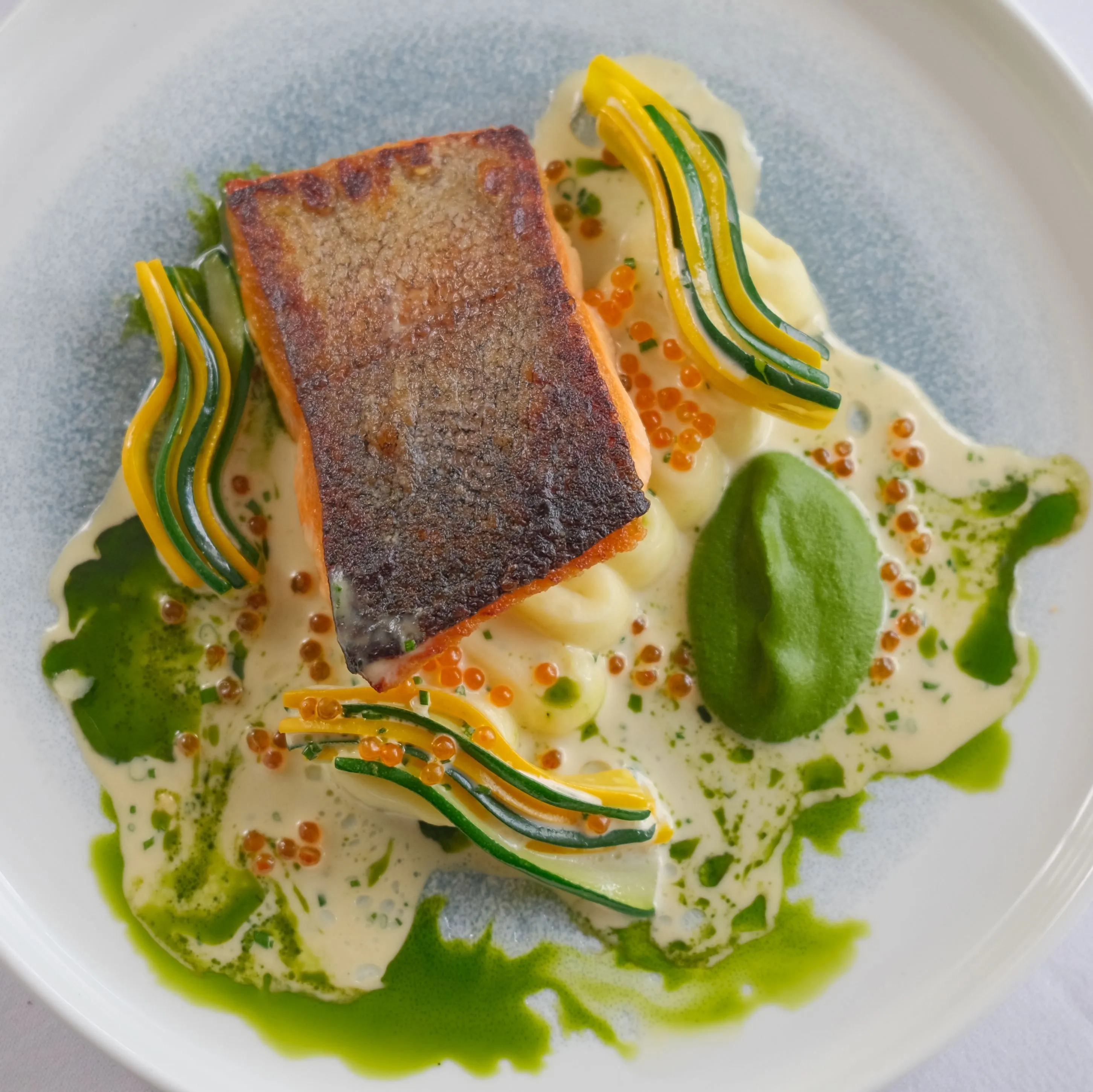 Pan-seared salmon fillet served on a white plate with a creamy sauce, green herb oil, fish roe and thin ribbons of courgette and yellow squash.