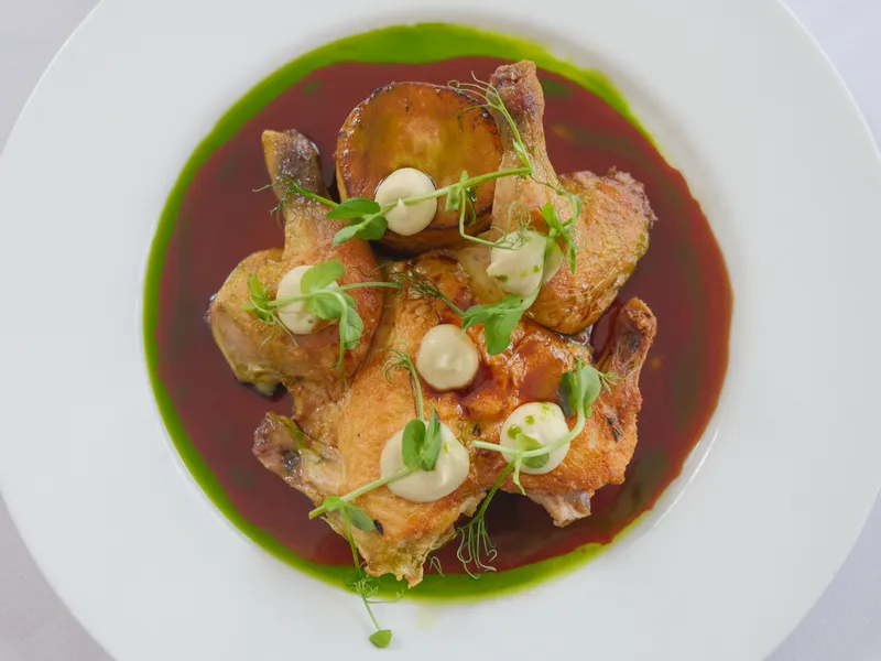 Roast chicken pieces plated with a rich brown sauce, green herb oil, a crisp potato round, and microgreens on a white plate