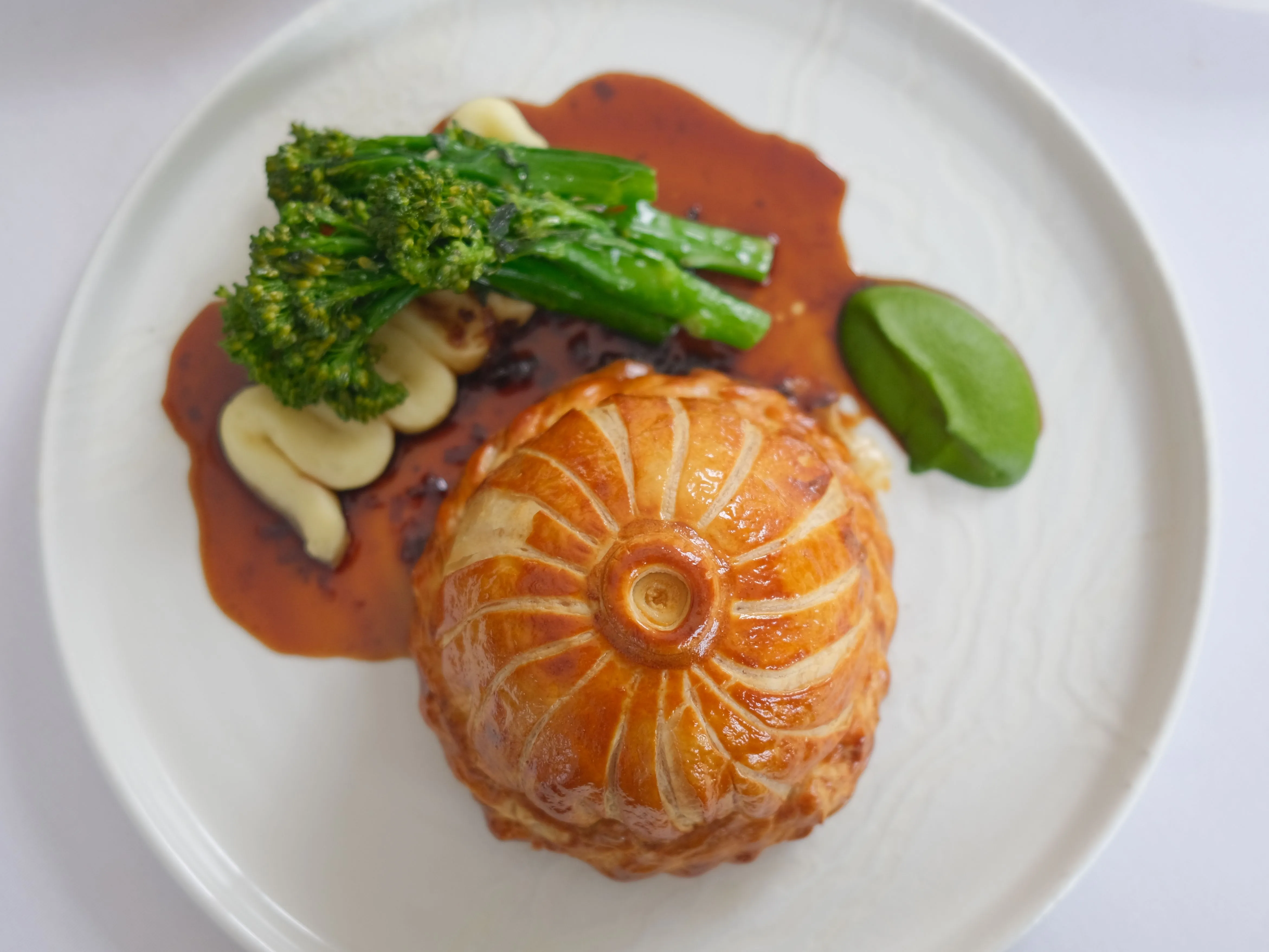 Golden puff pastry pie served with tenderstem broccoli, creamy mashed potato, red sauce and a green purée on a white plate