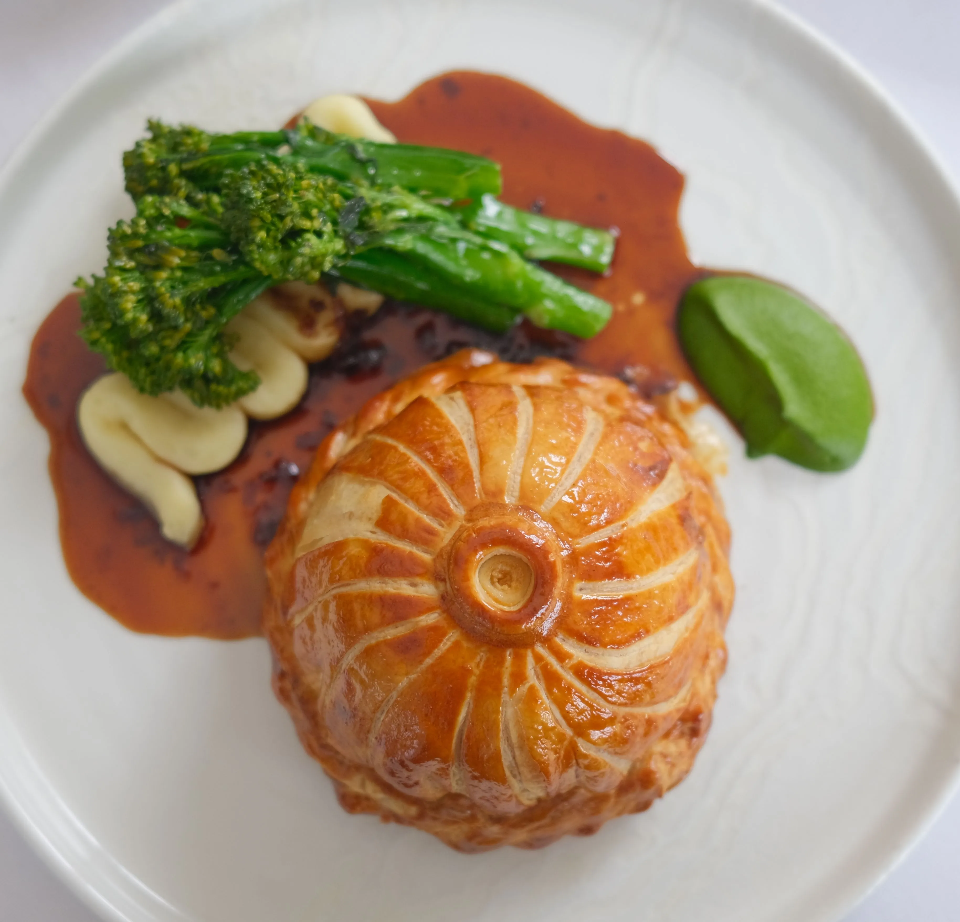 Golden puff pastry pie served with tenderstem broccoli, creamy mashed potato, red sauce and a green purée on a white plate