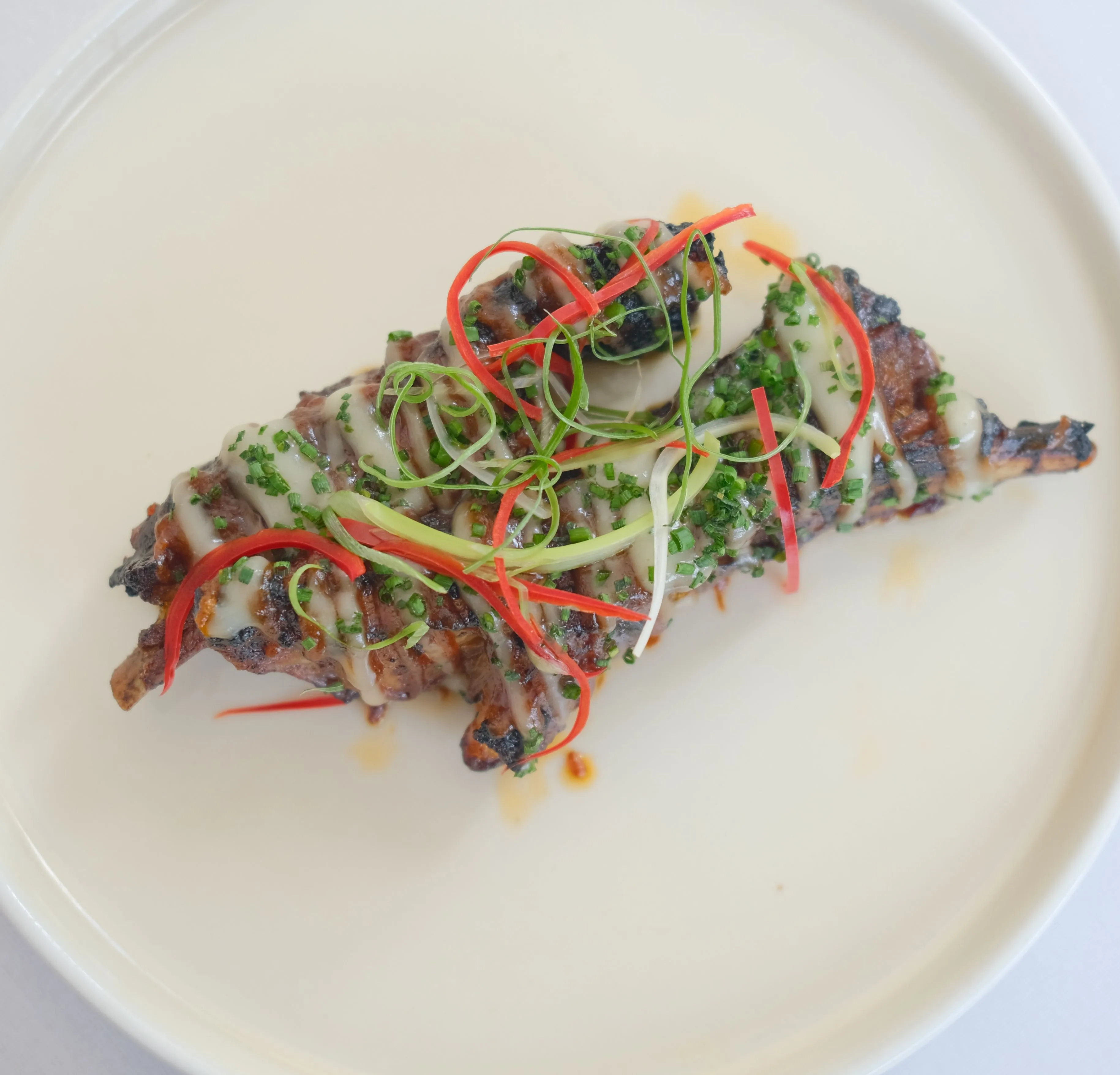 Grilled meat skewers on a white plate, drizzled with sauce and topped with spring onion and red chilli