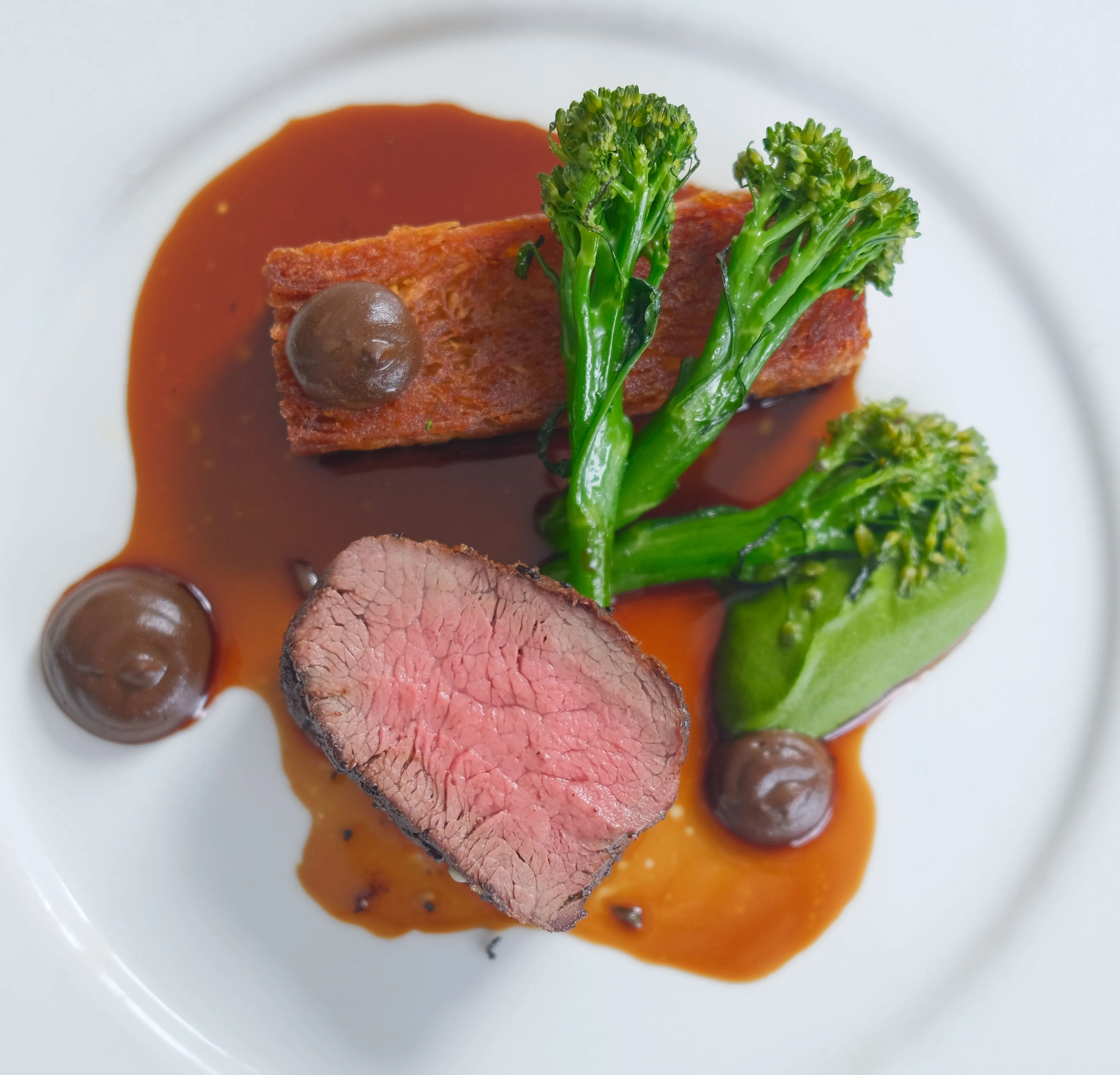 Plated fine dining dish with sliced pink beef, tenderstem broccoli, a crisp rectangular side and rich brown sauce on a white plate